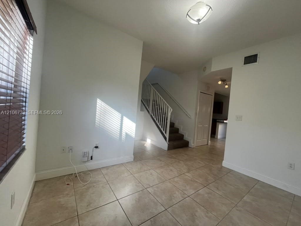 Interior space featuring large-format tile flooring, a central staircase with white railing, ceiling-mounted light fixtures, and windows with horizontal blinds