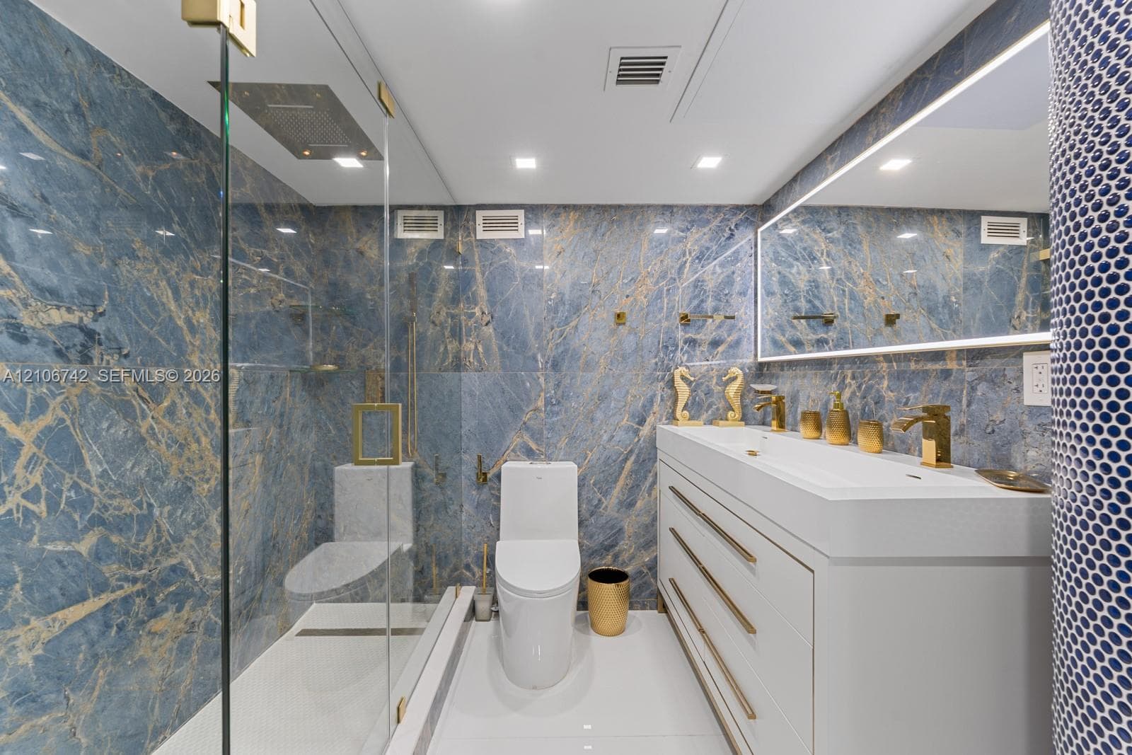 Full-wall blue-veined stone tile, walk-in glass-enclosed shower with a rainfall showerhead, white double-drawer vanity with gold hardware, integrated sink, and an illuminated mirror