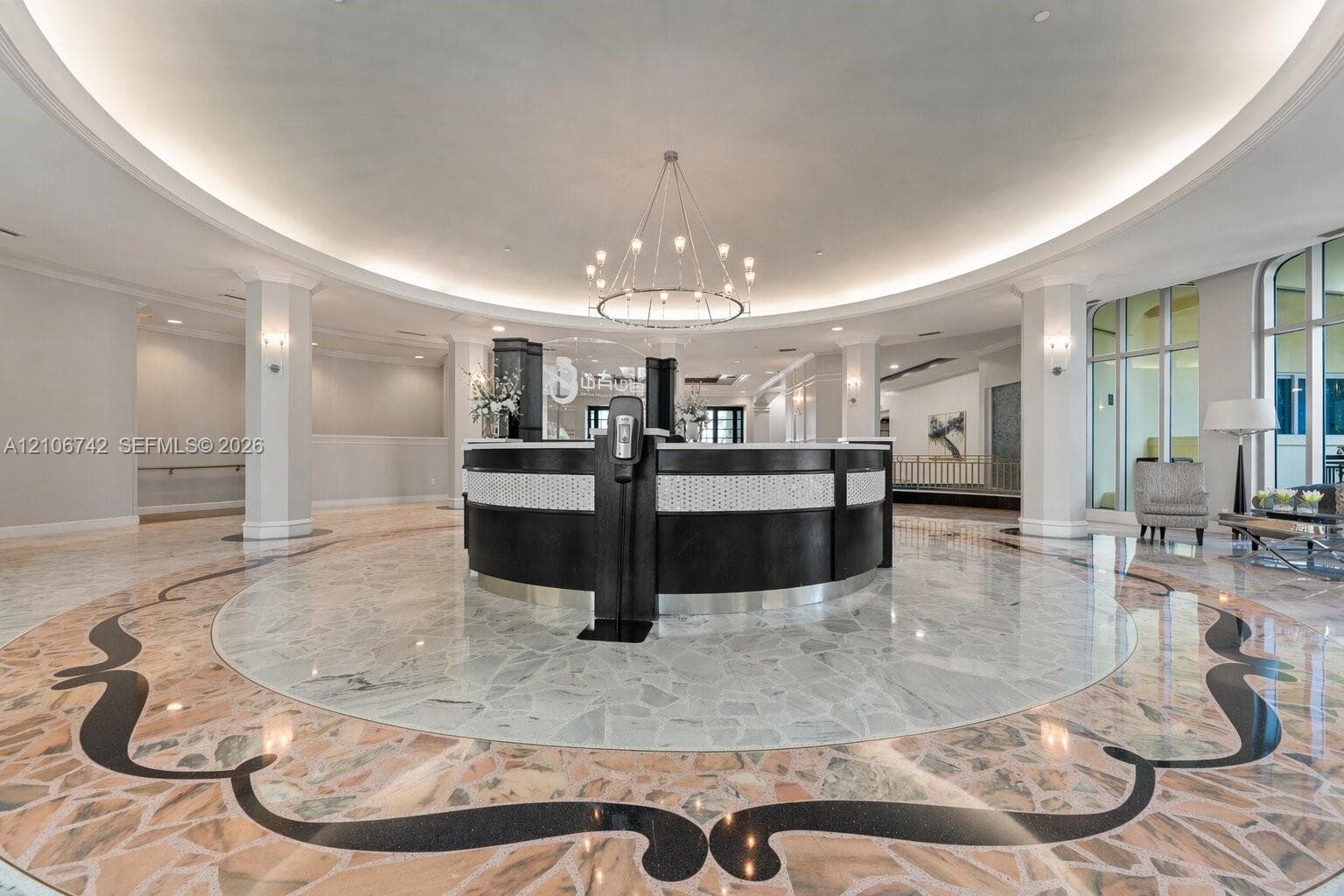 Grand foyer featuring decorative stone flooring with intricate inlays, a curved built-in reception desk, a central multi-arm chandelier, recessed ceiling cove lighting, and structural columns