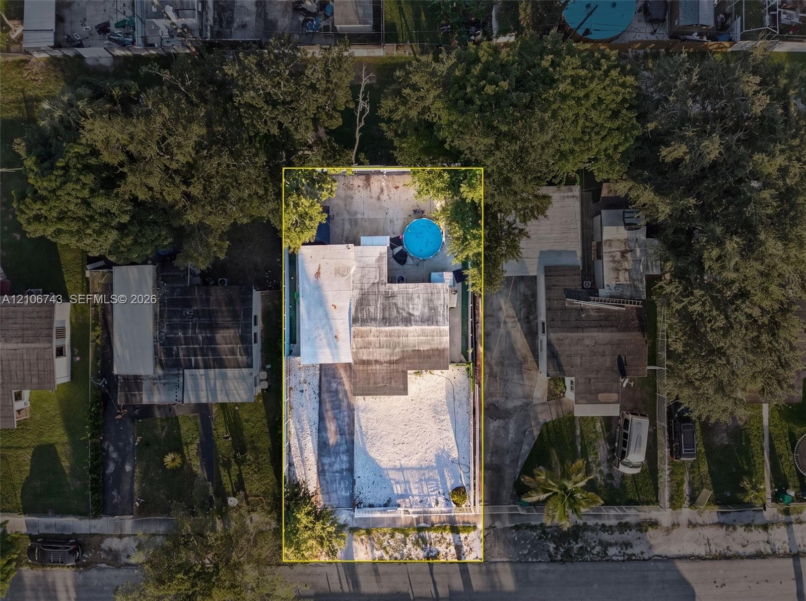Aerial perspective of a residential lot featuring a detached structure, circular above-ground pool, expansive gravel yard space, and mature perimeter trees
