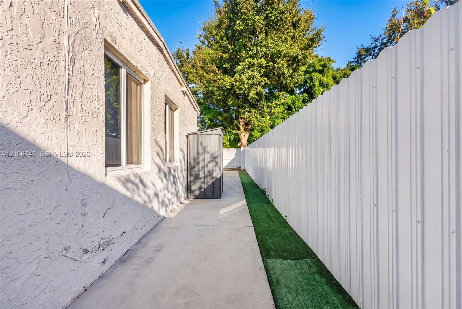 Exterior side yard featuring a concrete pathway, tall corrugated metal privacy fencing, and artificial turf bordering