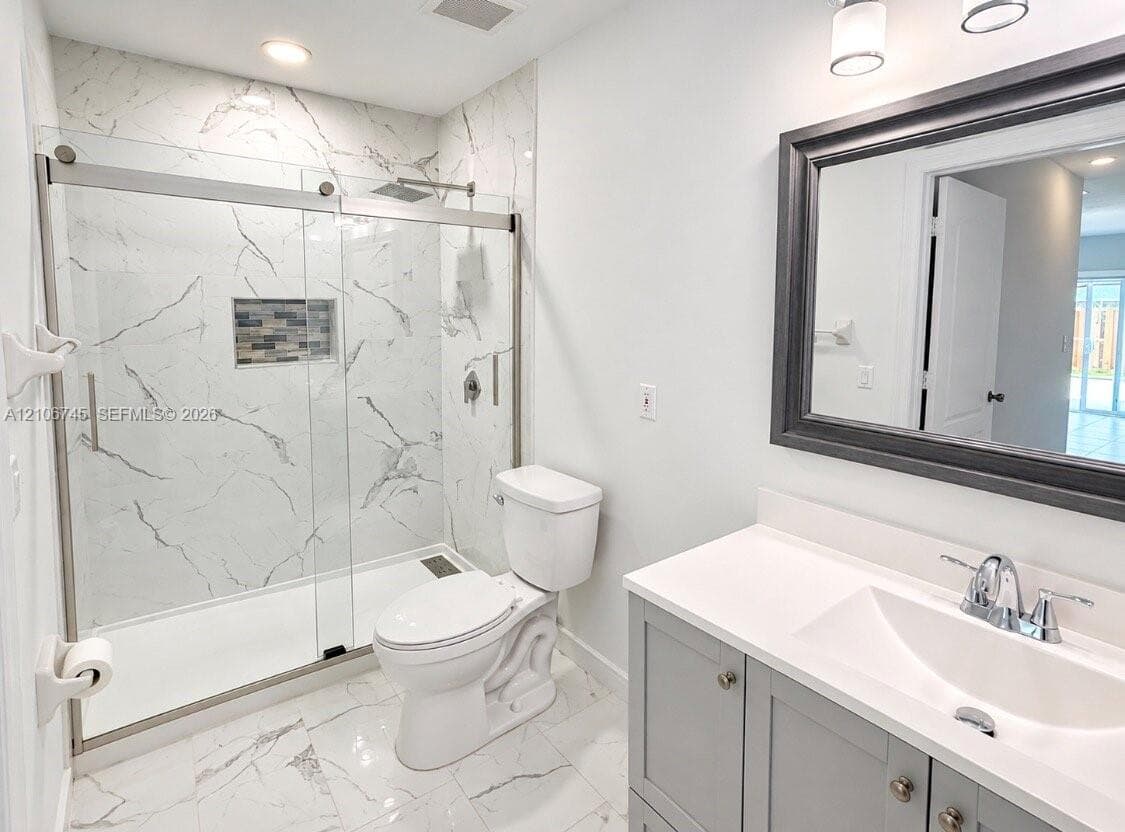 Walk-in shower with floor-to-ceiling stone-patterned tile, glass sliding door, recessed mosaic-tiled niche, gray vanity with white countertop, and framed mirror