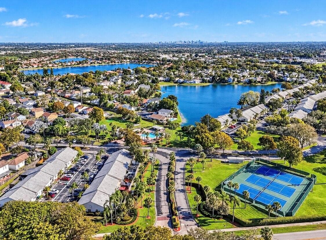 Aerial perspective of the community featuring an outdoor swimming pool, fenced tennis courts, manicured landscaping, and lakefront surroundings