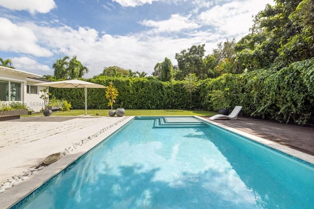 In-ground swimming pool with built-in steps, expansive wood-finish decking, light-colored stone patio surround, manicured lawn, and perimeter hedging