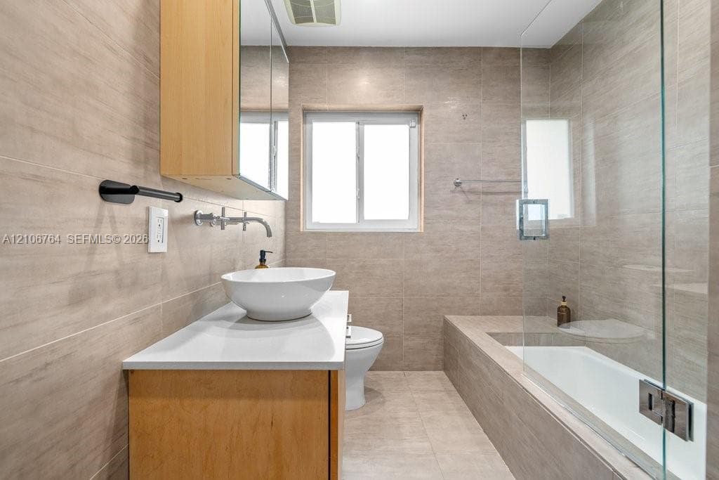 Bathroom featuring floor-to-ceiling neutral tile, custom wood-finish vanity with a white vessel sink, quartz countertop, wall-mounted faucet, and a glass-enclosed tub with shower