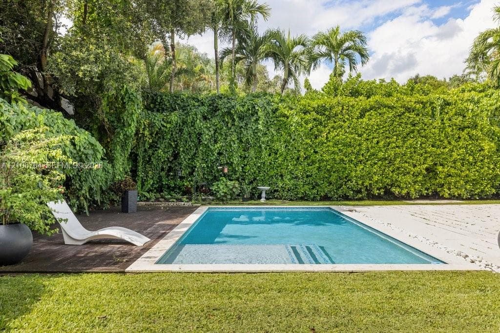 Rectangular swimming pool featuring a light-toned stone surround, wood-finish decking, and an expansive backdrop of manicured hedges and mature palm trees