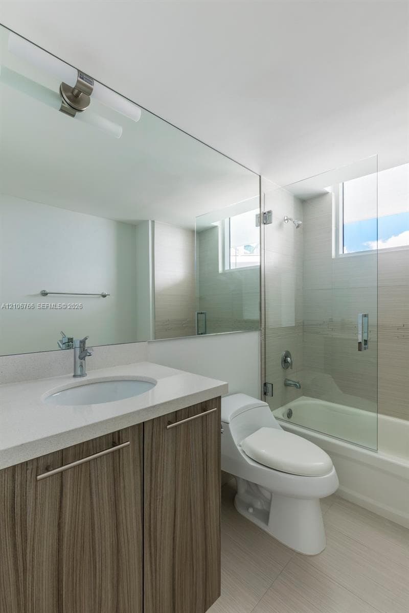Bathroom featuring a wood-finish vanity with a stone-surface countertop, an integrated sink, a full-width wall mirror, and a glass-enclosed tub with tiled surround