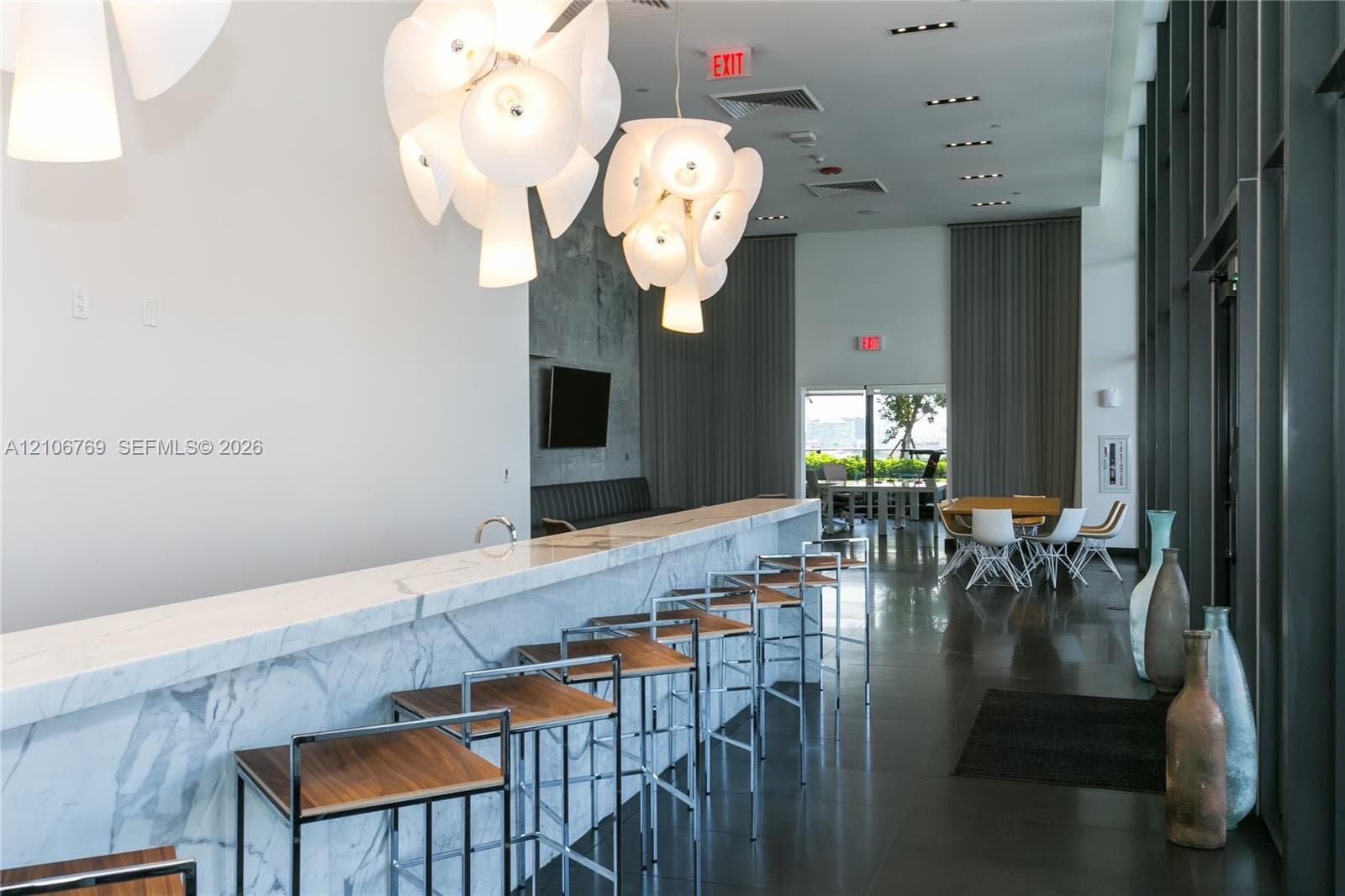 Full-height floor-to-ceiling windows, stone-finish bar with integrated sink, polished tile flooring, recessed ceiling lighting, and sculptural pendant chandeliers