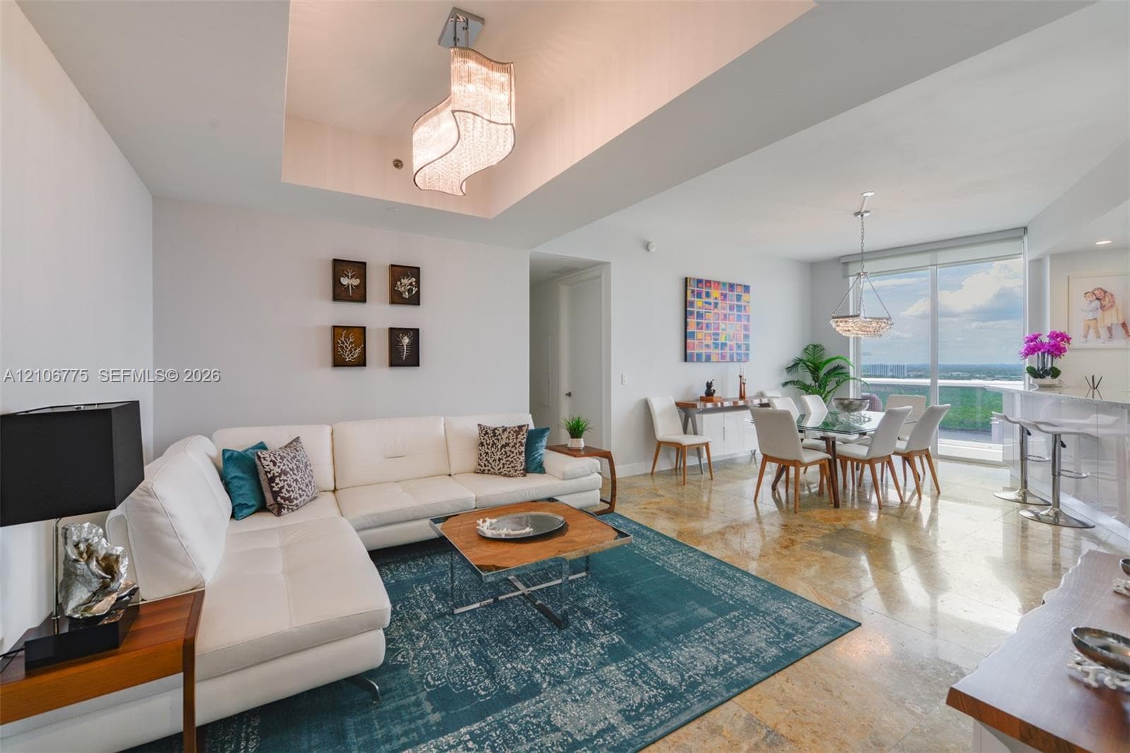 Open-concept interior featuring natural stone flooring, tray ceiling with a contemporary tiered chandelier, floor-to-ceiling windows, and built-in breakfast bar