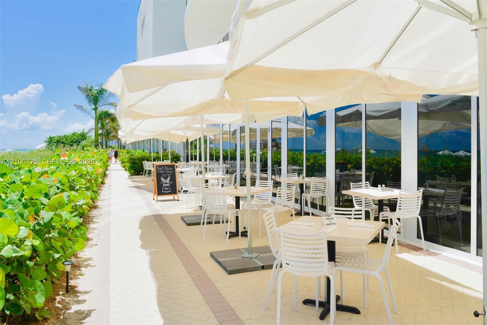 Outdoor paved patio featuring large-format umbrellas, glass exterior wall panels, and manicured perimeter landscaping