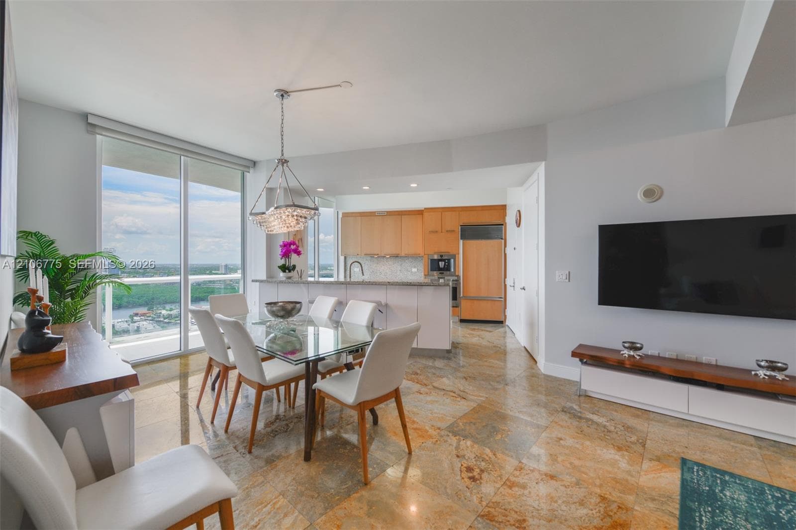 Open-concept interior featuring natural stone flooring, floor-to-ceiling windows, built-in wooden cabinetry, recessed lighting, and a pendant chandelier