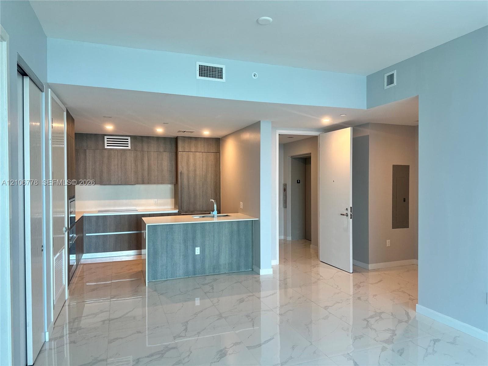 Modern open-concept living area featuring polished stone flooring, a central kitchen island with wood-finish cabinetry, stainless steel appliances, recessed ceiling lighting, and abundant natural light from expansive glass sliding doors