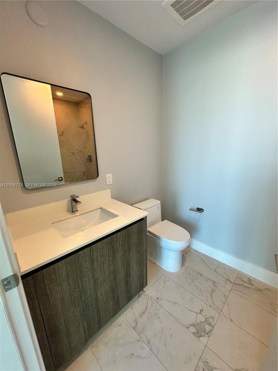 Modern vanity with a rectangular sink, stone-finish countertop, and wood-texture cabinetry, complemented by large-format tile flooring and a wall-mounted mirror with a black frame