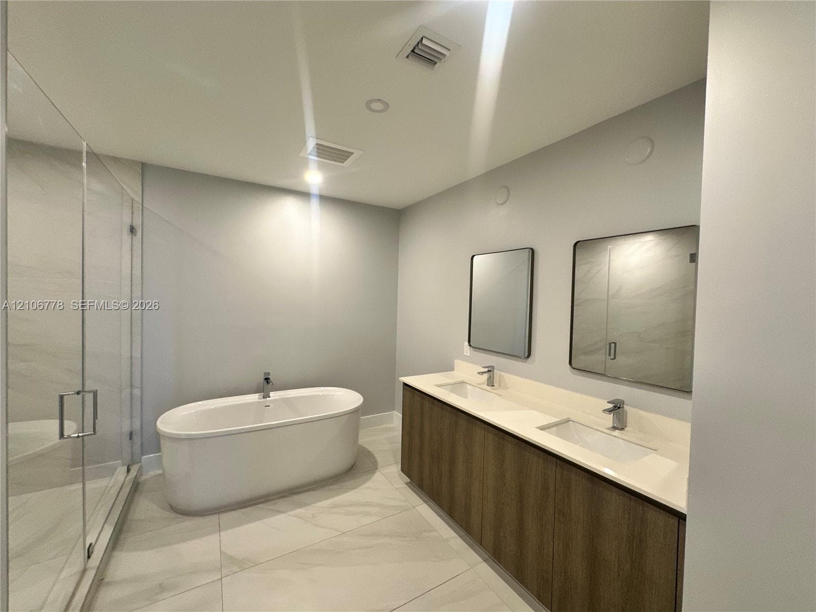 Dual-sink vanity with wood-finish cabinetry, quartz countertop, freestanding soaking tub, and glass-enclosed walk-in shower with tile surround