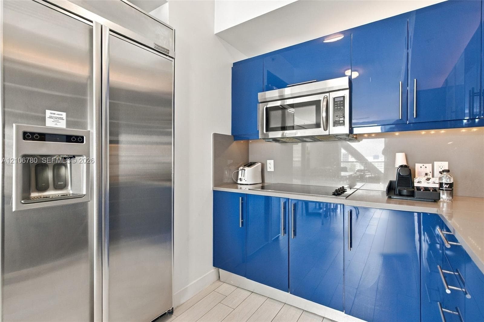 Modern kitchen featuring high-gloss blue cabinetry, stone-finish countertops, a glass-surface cooktop, a stainless steel over-the-range microwave, and a full-size stainless steel refrigerator