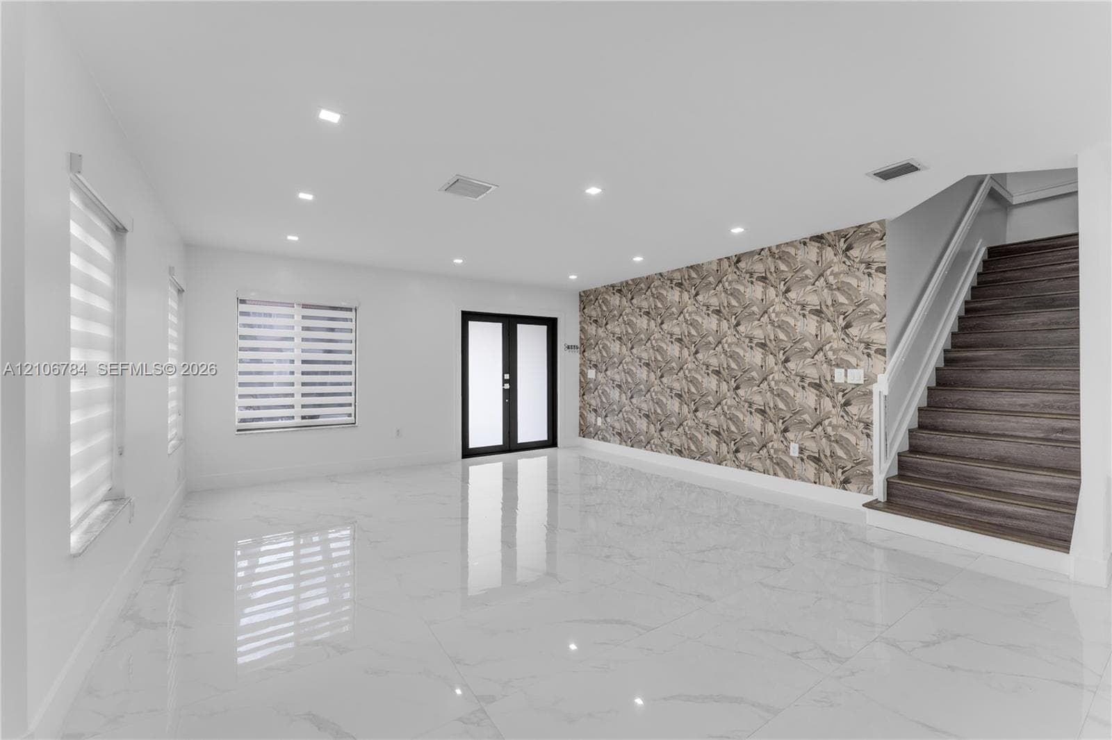 Open-concept room featuring large-format tile flooring, recessed lighting, double-door entryway, patterned accent wall, and staircase with wood-finish treads