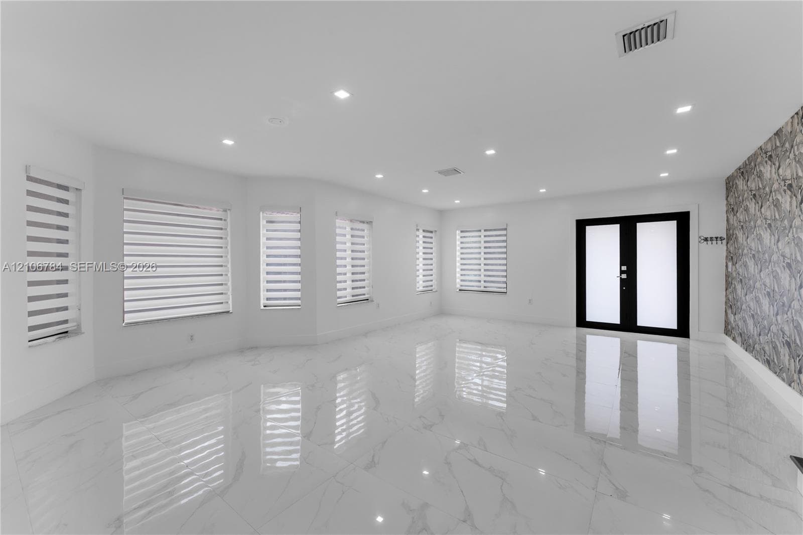 Open-concept area featuring polished porcelain tile flooring, recessed lighting, textured stone accent wall, dual-entry doors with frosted glass panels, and multiple windows with dual-layer zebra shades