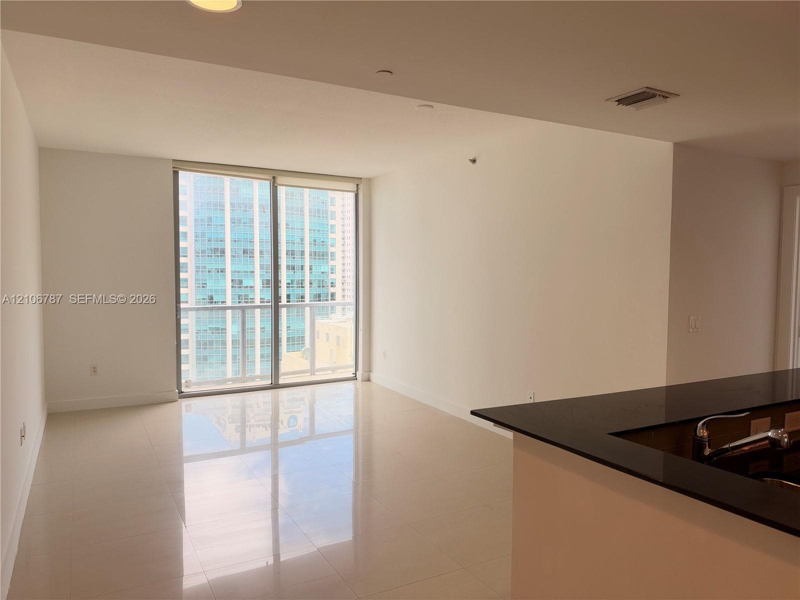 Floor-to-ceiling sliding glass doors with balcony access, high-gloss tile flooring, and kitchen featuring a dark stone-finish countertop