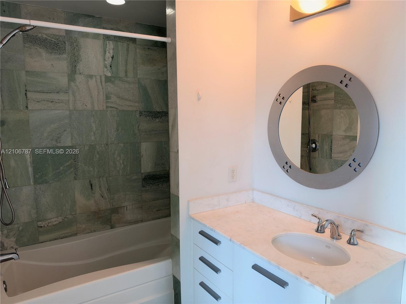 Bathroom featuring a vanity with stone-finish countertop, undermount sink, circular wall-mounted mirror, and tile-surround bathtub