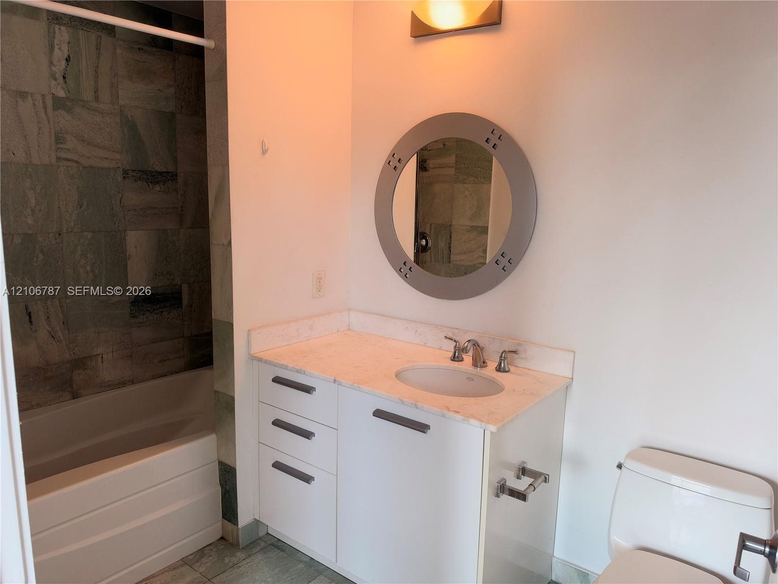 Bathroom featuring a stone-topped vanity with white cabinetry, integrated sink with chrome faucet, circular wall-mounted mirror, and tile surround tub area