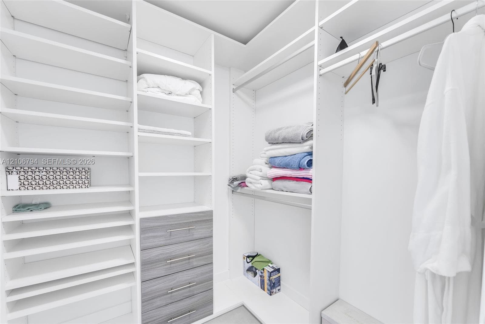 Walk-in closet featuring floor-to-ceiling white shelving, integrated hanging rods, and a four-drawer dresser unit with grey wood-finish drawer fronts and silver-toned pulls