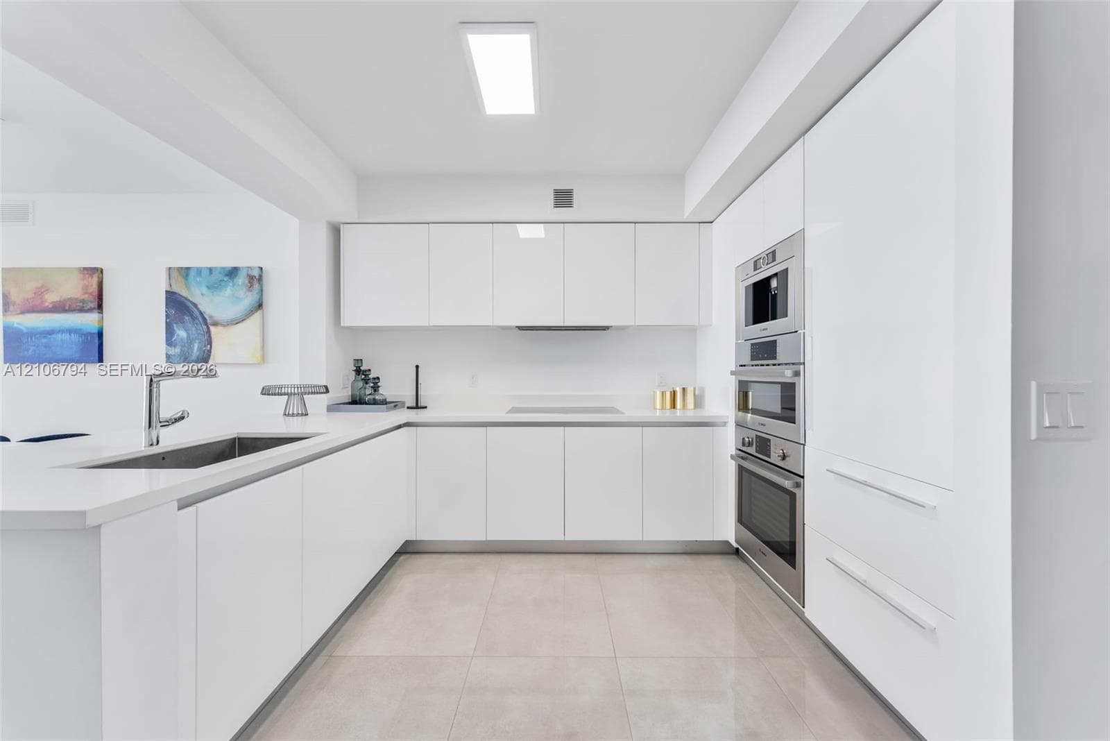Modern kitchen featuring white flat-panel cabinetry, light-toned tile flooring, integrated stainless steel appliances, and solid-surface countertops with an undermount sink