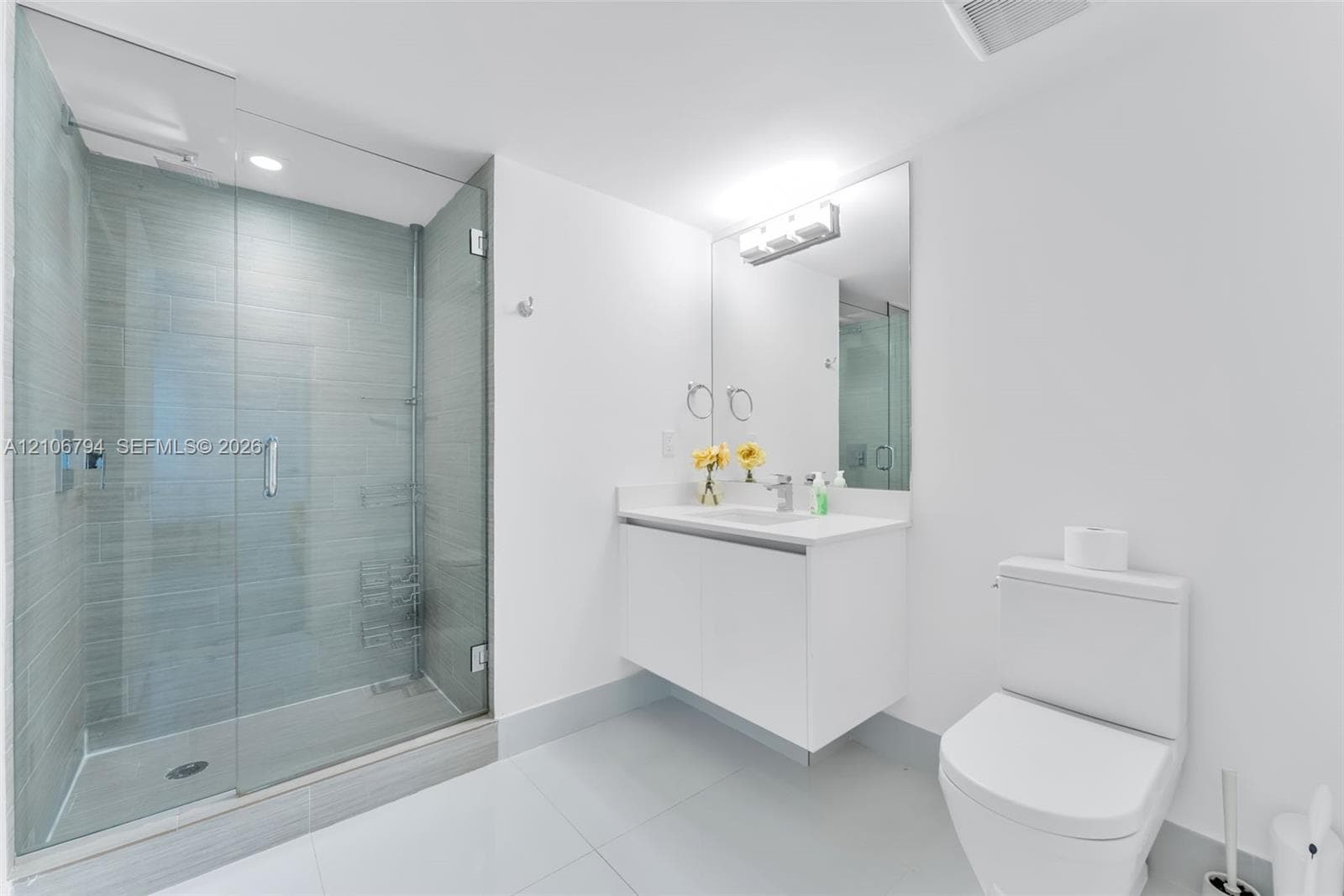 Walk-in shower with horizontal tile surround and glass door, wall-mounted white vanity, solid-surface countertop with integrated sink, and large-format tile flooring