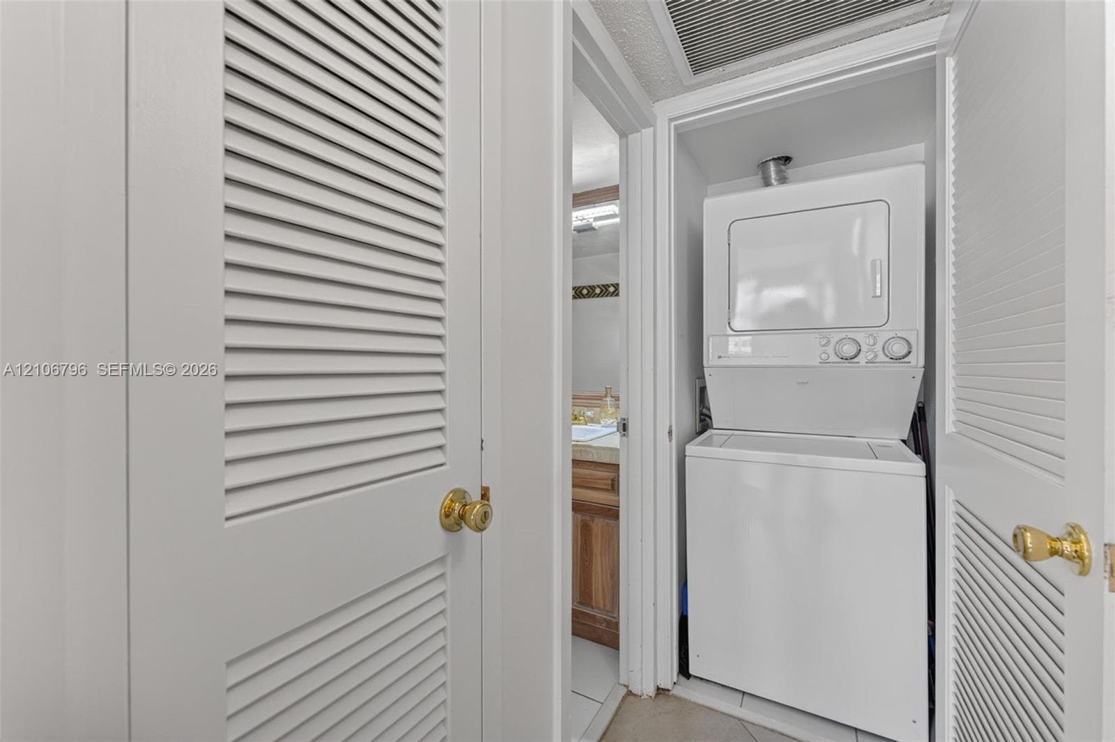 Stacked laundry unit featuring white cabinetry, louvered closet doors with brass-finish hardware, and tiled flooring