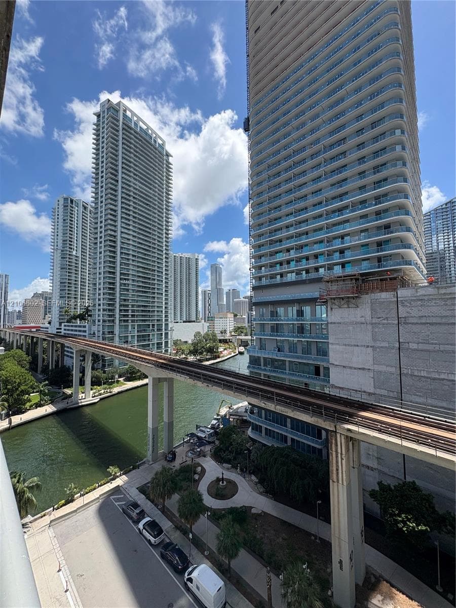 Urban high-rise setting featuring high-density residential towers, an elevated transit rail system, a waterway, and concrete-paved walkways with established palm trees