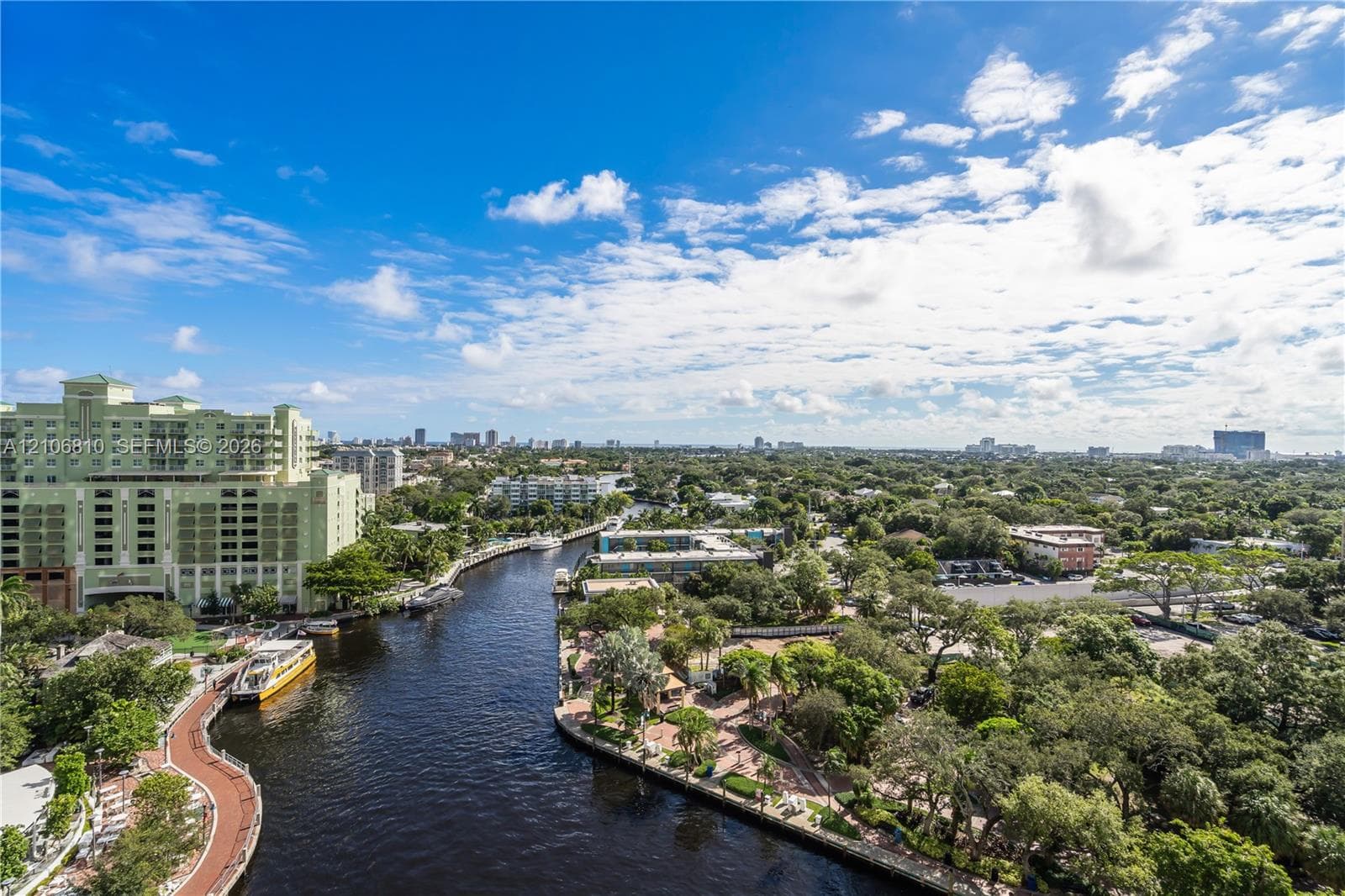 Elevated aerial perspective featuring a winding waterway, paved pedestrian paths, mature tropical landscaping, and a broad cityscape horizon