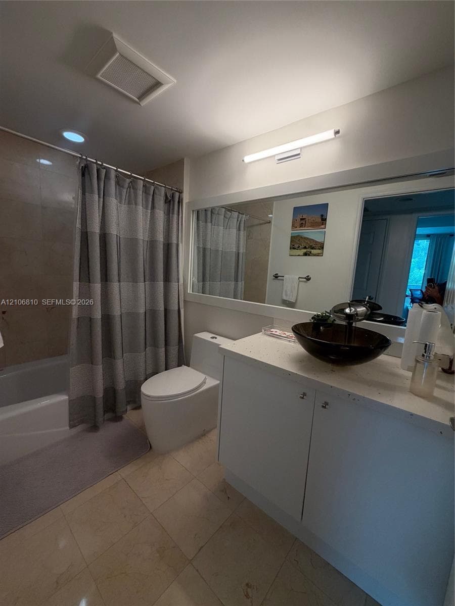 Bathroom featuring stone-finish floor tiling, a white vanity with a light-colored solid surface countertop, a dark glass vessel sink, and a large wall-mounted mirror