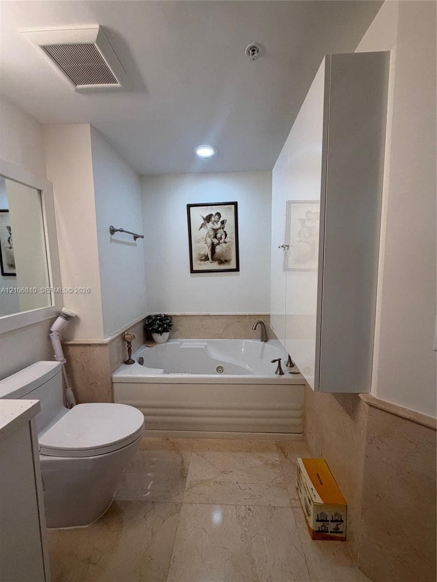 Bathroom featuring a built-in jetted soaking tub, stone tile flooring, matching stone wainscoting, and a white high-gloss wall-mounted cabinet