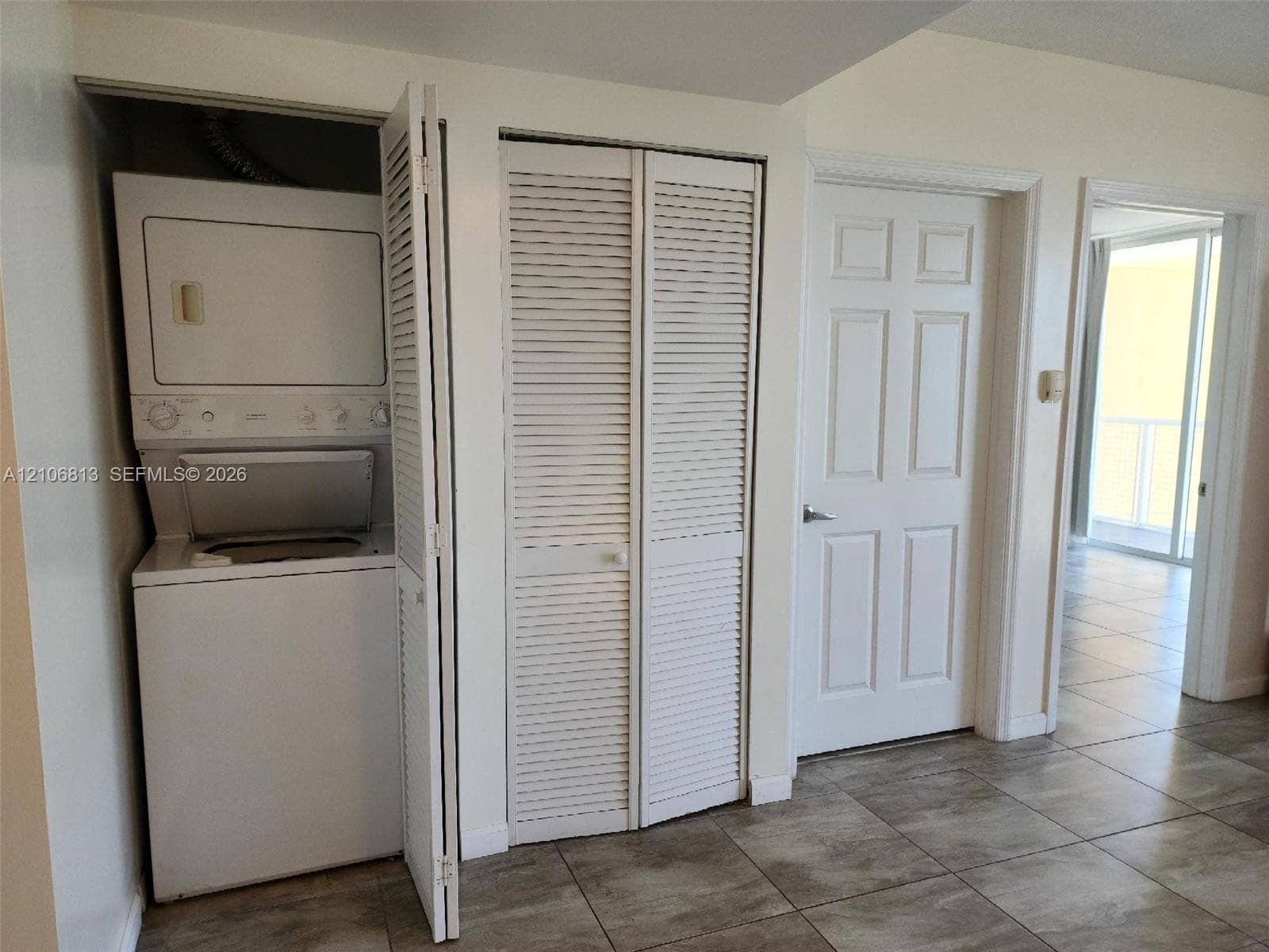 Stacked laundry unit, white louvered closet doors, and ceramic tile flooring