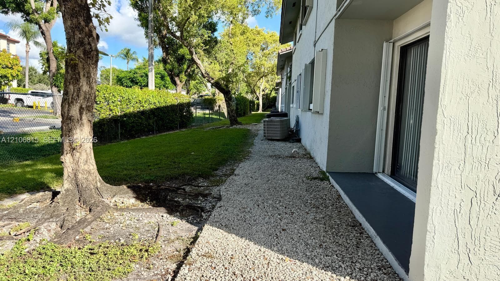 Exterior side yard featuring a gravel pathway, concrete foundation, manicured lawn, and mature trees