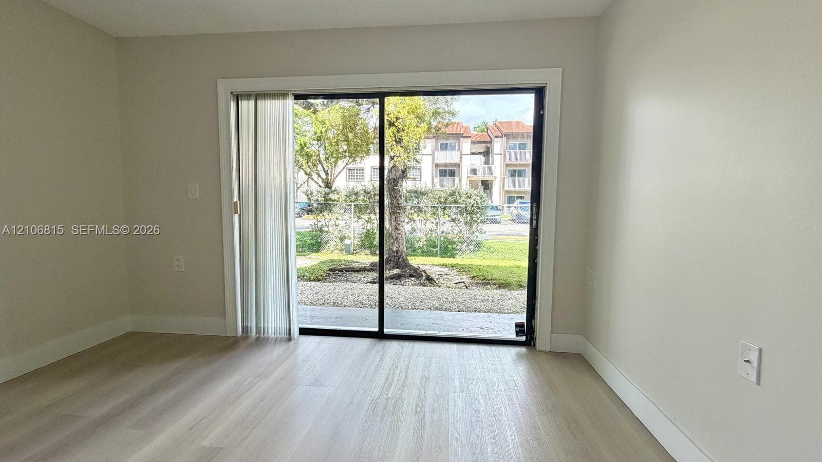Neutral interior space featuring light-toned wood-finish flooring, smooth wall finishes, and a black-frame sliding glass door with vertical blinds