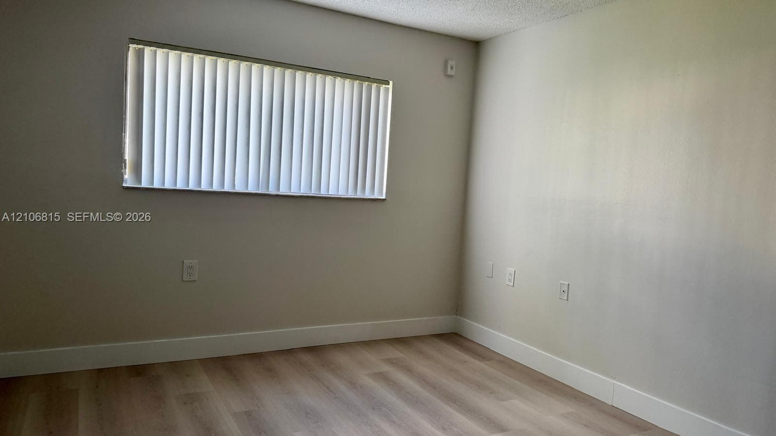 Neutral-toned interior space featuring light wood-finish flooring, smooth wall finishes, and a window equipped with vertical blinds