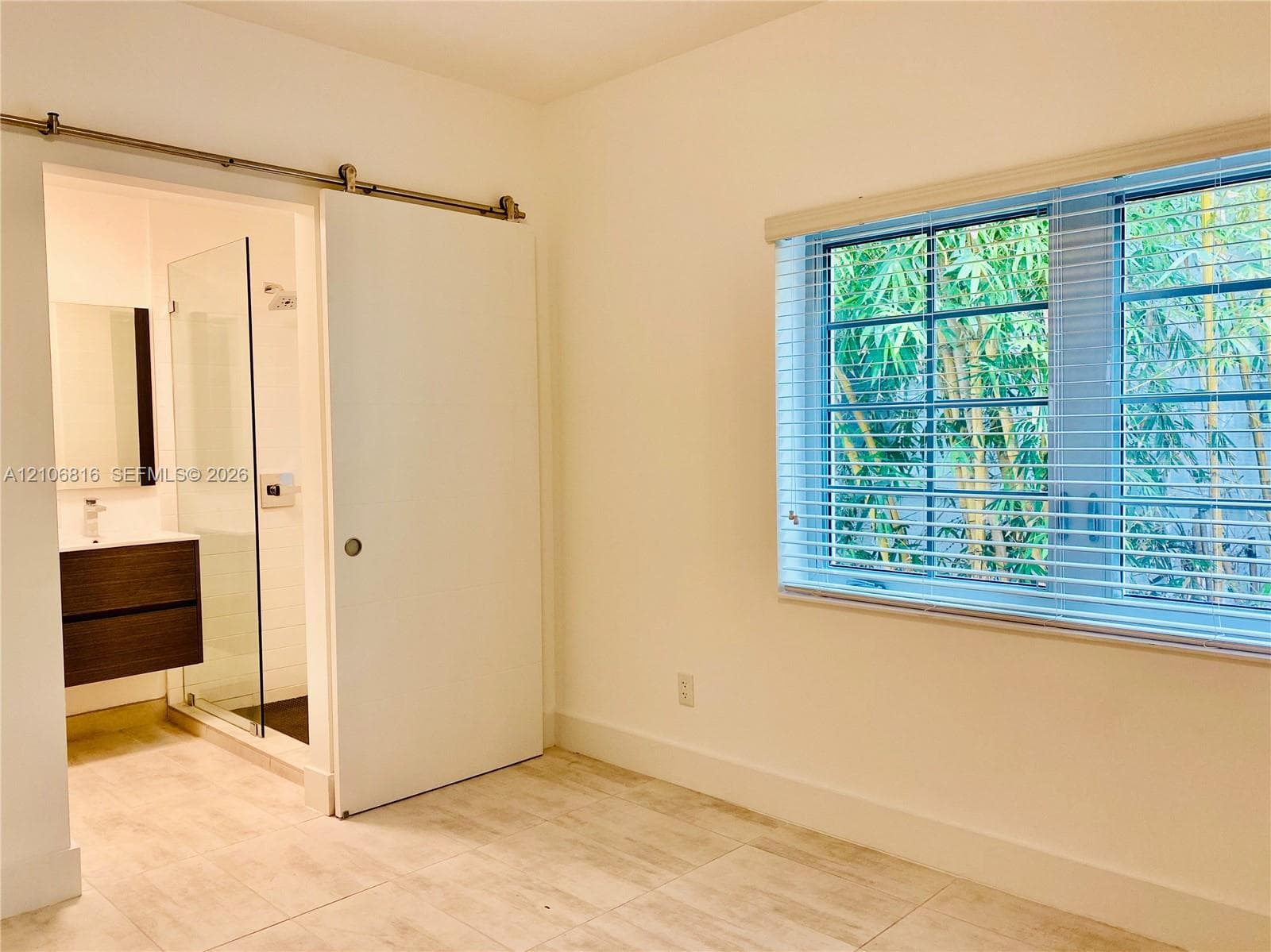 Interior space featuring tile flooring, sliding barn door with metal hardware, multi-pane window with horizontal blinds, and attached bathroom with floating vanity and glass-enclosed shower