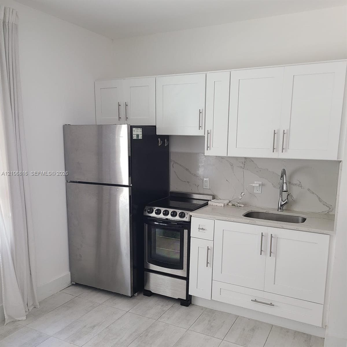 Kitchen area featuring white shaker-style cabinetry, a stone-patterned backsplash, stainless steel refrigerator and range, and light-toned tile flooring