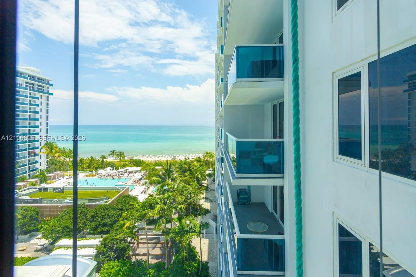 High-rise building exterior featuring private balconies with glass railings and reflective windows, overlooking the ocean, coastline, and tropical landscaping