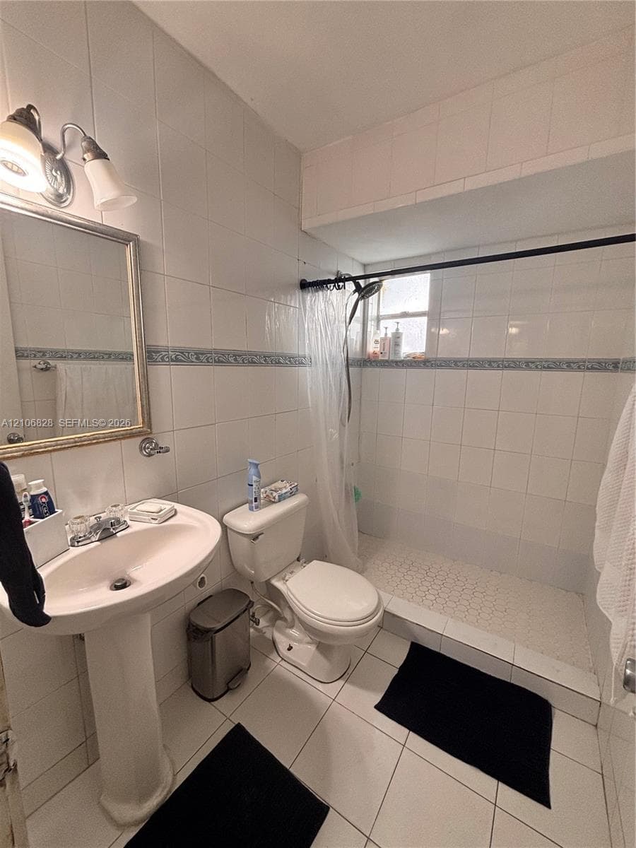 Full-bathroom featuring a white pedestal sink, tiled walk-in shower with integrated shelving, wall-mounted light fixture, and ceramic tile flooring