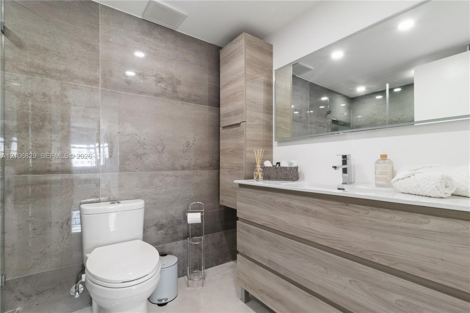 Bathroom featuring large-format gray tiled walls, vanity with wood-finish cabinetry, integrated countertop sink, tall built-in storage cabinet, and wall-to-wall mirror