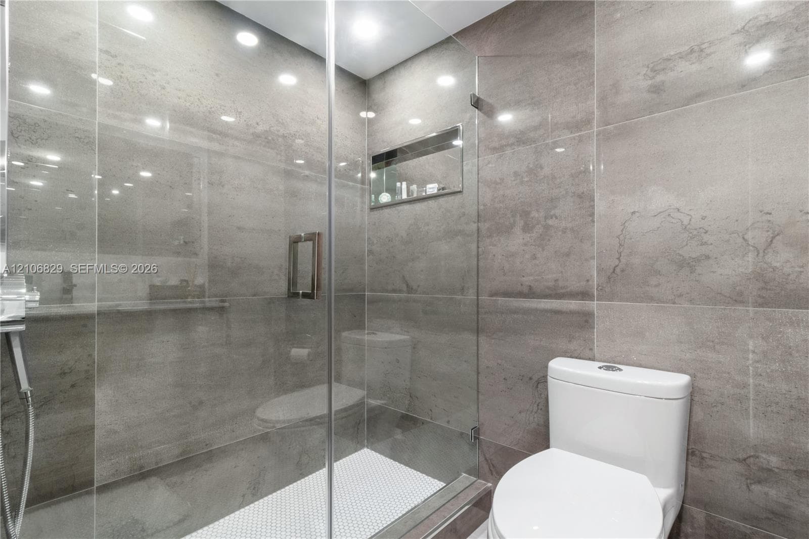 Walk-in shower featuring large-format grey tile, a frameless glass enclosure, a built-in recessed shelf, and white mosaic floor tile, accompanied by a dual-flush toilet