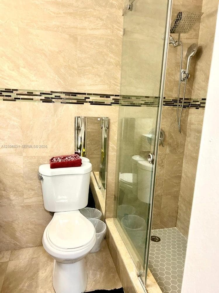 Bathroom featuring floor-to-ceiling stone-finish tile, glass-enclosed shower with a rainfall showerhead and handheld sprayer, and an integrated glass mosaic accent border