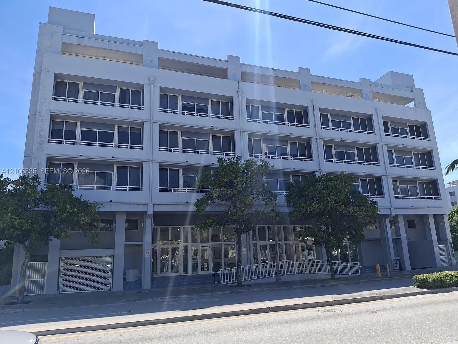 Multi-story concrete structure featuring glass-paneled balconies, floor-to-ceiling storefront windows on the ground level, and exterior privacy shutters