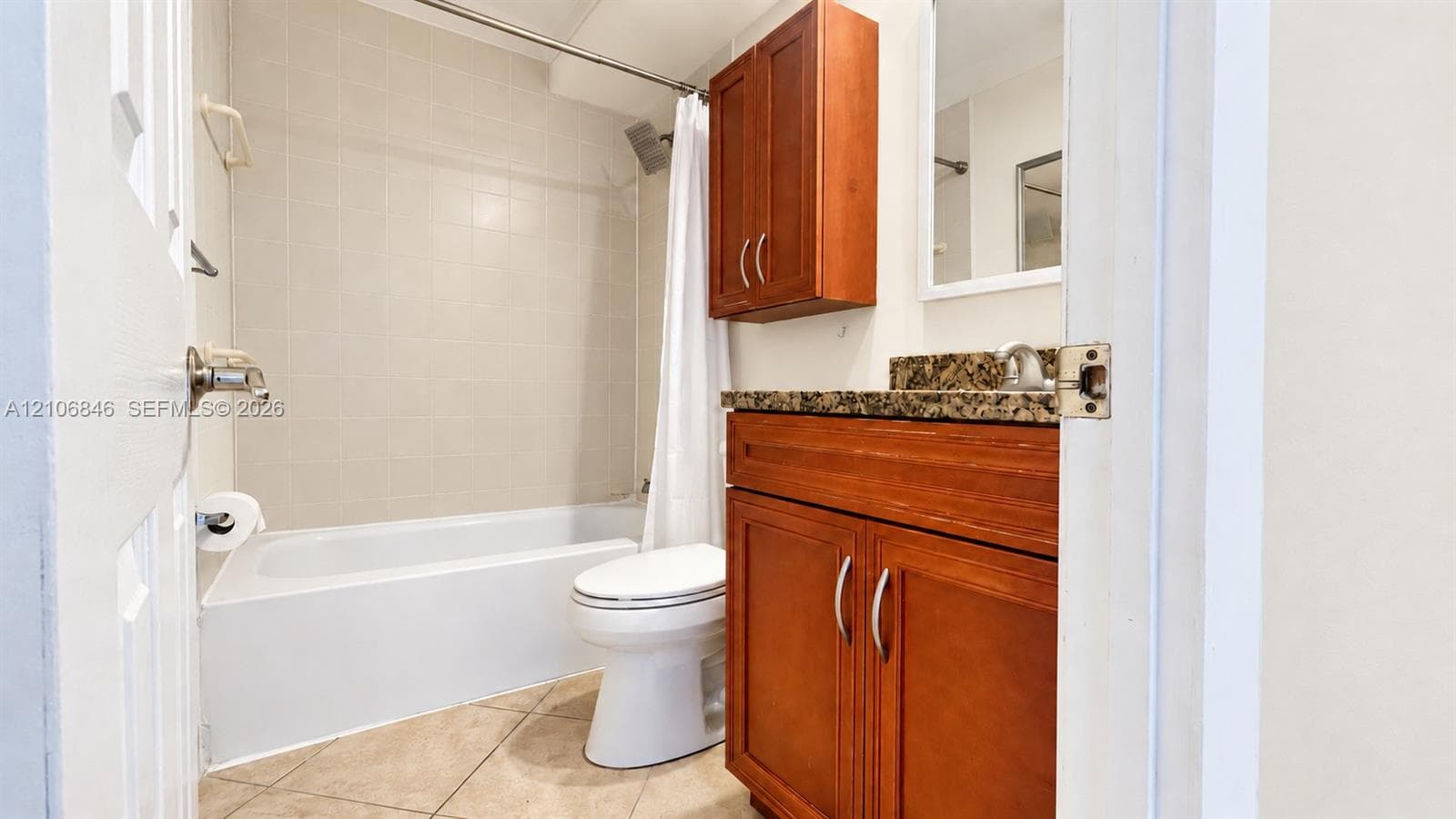 Bathroom featuring a wood-finish vanity cabinet, stone-surface countertop, integrated wall-mounted storage cabinet, tiled tub enclosure, and neutral floor tile