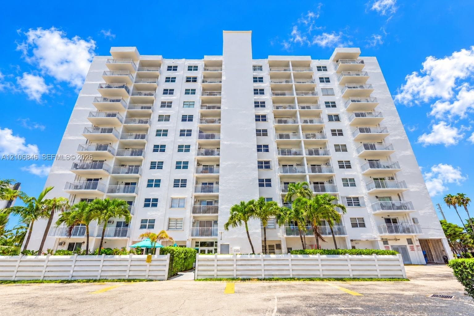 Multi-story residential building featuring a white facade, private balconies for each unit, glass-panel railings, and perimeter privacy fencing