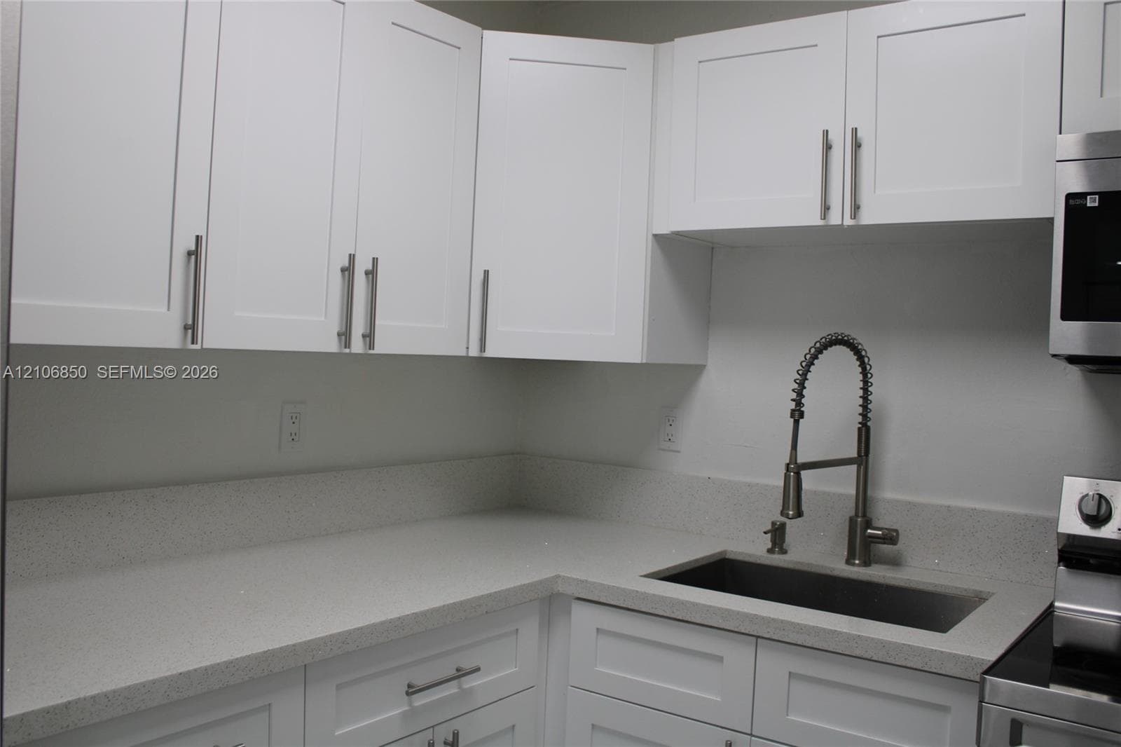 White Shaker-style cabinetry with polished metal hardware, light-toned solid surface countertops, a stainless steel undermount sink, and a high-arch pull-down faucet