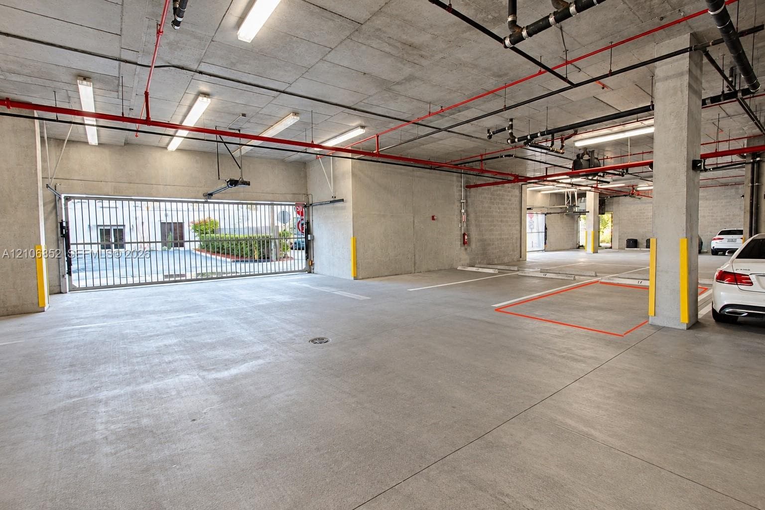 Covered parking area featuring concrete flooring, structural concrete columns, overhead industrial lighting, and a gated perimeter entrance