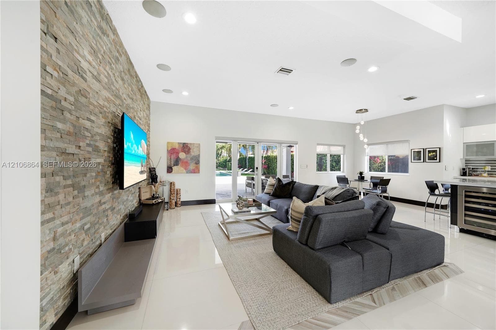 Open-concept living space featuring high ceilings with recessed lighting, large-format tile flooring, a floor-to-ceiling stone-veneer accent wall, and a kitchen equipped with a stainless steel range hood and built-in wine refrigeration
