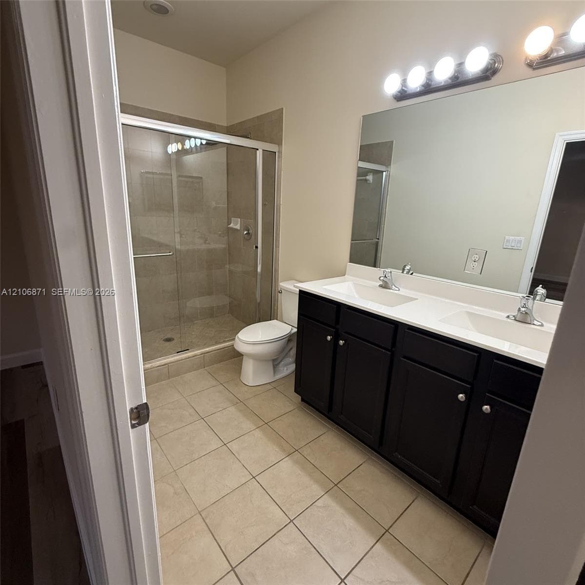 Bathroom featuring a double-sink vanity with dark-finish cabinetry, a solid-surface countertop, a large wall-mounted mirror, and a walk-in glass-enclosed shower with tile surround
